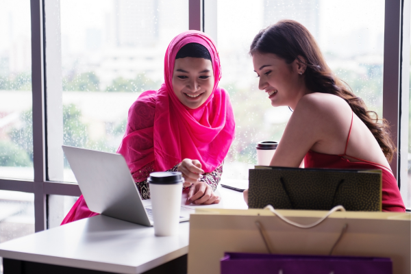 Two young females from different cultures are looking at a computer and smiling. An Islamic female is dressed in a pink Hijab. 