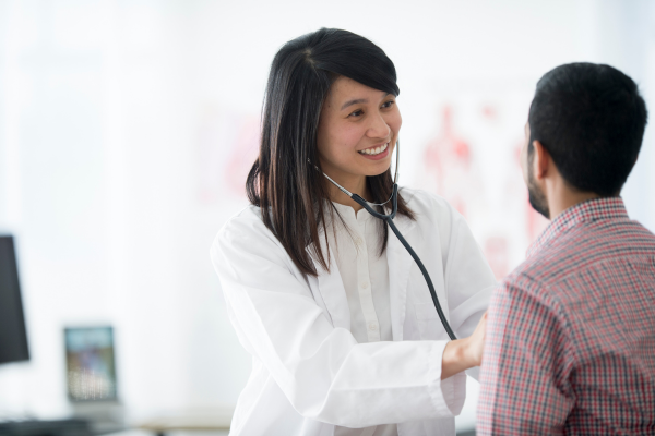 A female doctor is using a stethoscope on a male patient. Getting a provincial health card is one of the first steps to take when settling in Canada.
