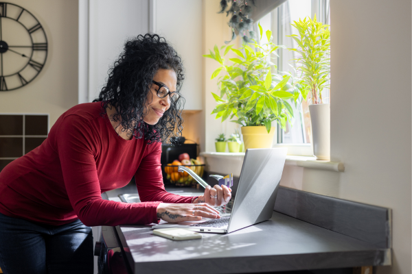 A female newcomer is standing in her kitchen and paying a utility bill online. Paying bills on time is a smart way Canada to manage your money and boost your credit score in Canada. 