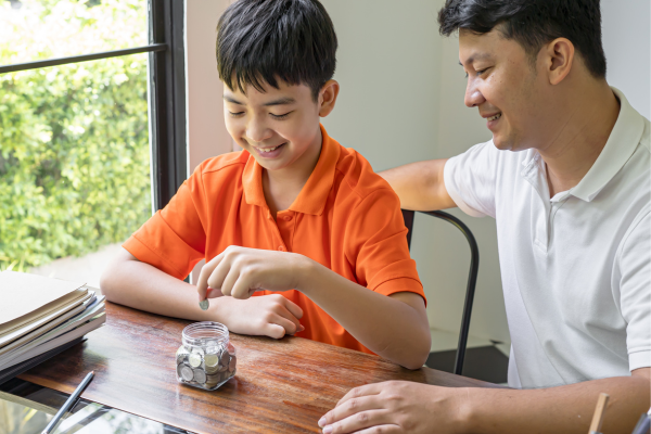 An Asian man is sitting with his young son as he deposits money into a savings jar. Financial literacy is vital for newcomers to achieve future goals. 