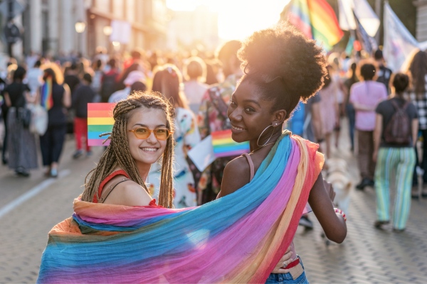 Two women with a pride flag draped around them while they are celebrating at a pride parade.