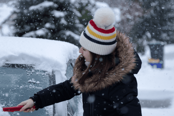 A woman brushing snow off of her car to prepare to drive in the winter.