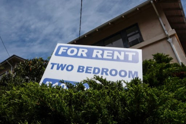 A sign advertising a 2-bedroom apartment for rent is placed in the hedges in front of an apartment building.