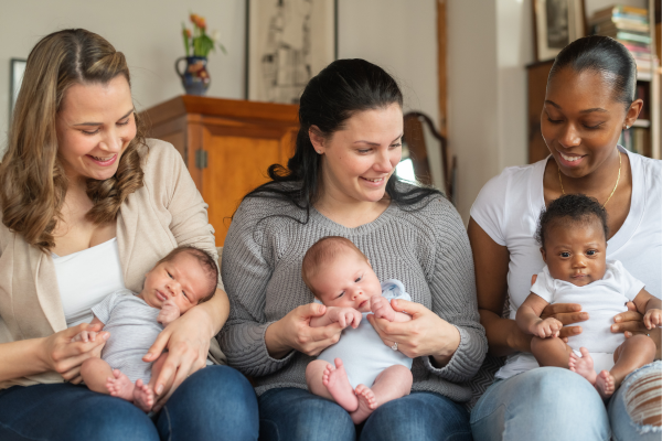 Three young mothers are sitting on a sofa with infant babies on their laps. They are celebrating their first Mother's Day.