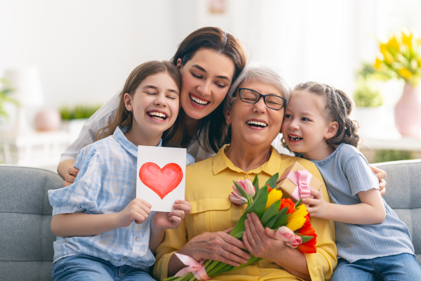 Young children are happily presenting cards, gifts, and flowers to their mom and grandmother for mom's day. 
