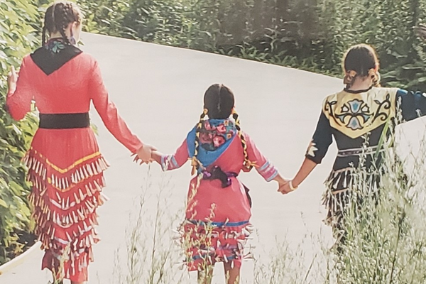 A picture of three young Indigenous children walking along a path, holding hands and wearing wearing traditional regalia. The photo represents truth and reconciliation