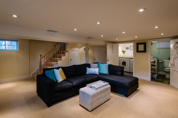 A walk-up basement apartment is mostly located below ground and offers less natural light. A sofa is located in the living room and you can see the entrance to the bathroom and laundry rooms. 