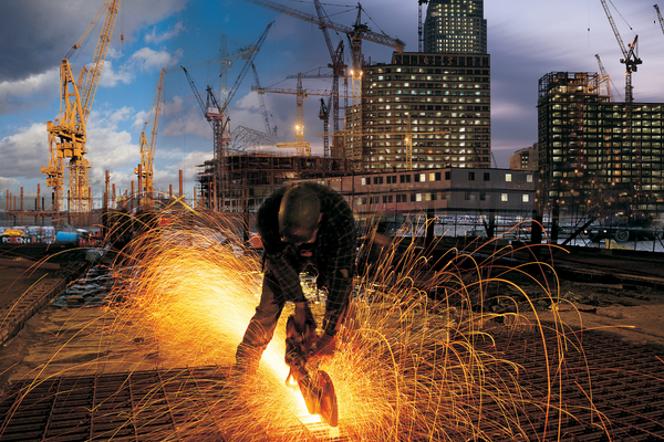Welder working outdoors on a construction site with cranes and building towers in the background
