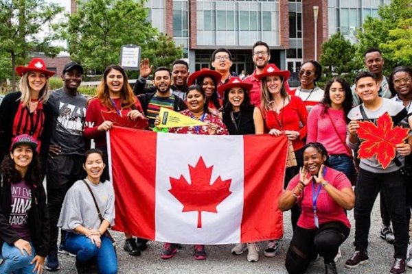 A happy group of students are holding a flag of Canada while standing in front of student housing. Tenant insurance for international students is vital.