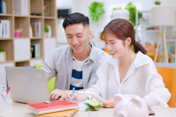 A young Asian couple discussing finances and money management while working on a laptop at home. Newcomers must carefully manage money and expenses in their first few months until their finances stabilize.