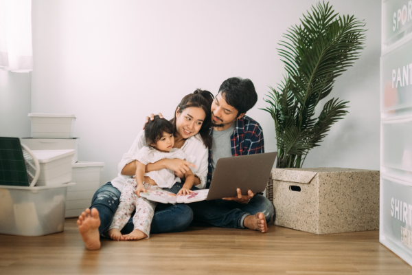 A young Asian man is seated on the floor with his wife and baby in their new rental apartment. 