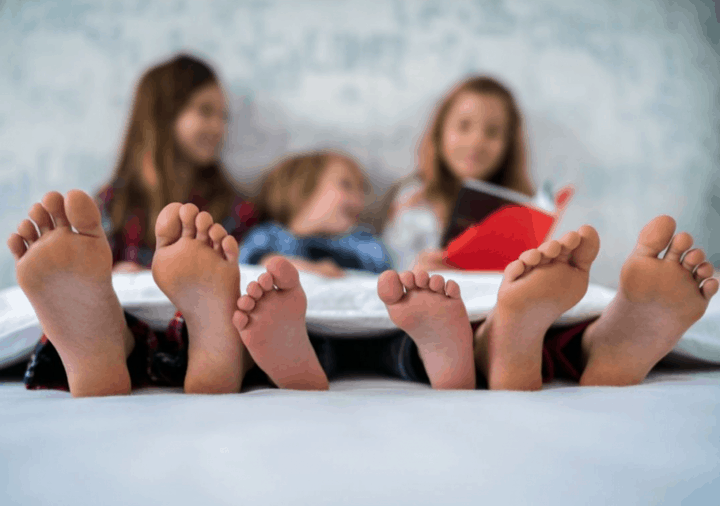 Three young children lying on a bed and reading a book. Their faces are blurred and their feet are clearly visible. Rules about children sharing bedrooms.